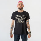 Young person with pink hair and tattoos wearing the Support Your Local Lager Maker T-shirt, showcasing their allegiance to local breweries with bold script on a black tee.