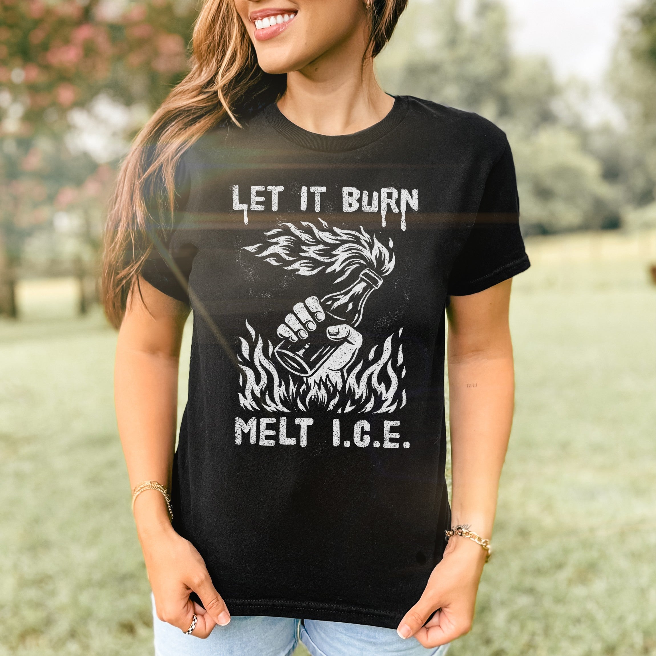 Woman wearing Let It Burn / Melt ICE Women's Baby Tee featuring Molotov cocktail graphic and text. The fitted black tee complements her casual jeans in a grassy, tree-lined setting.