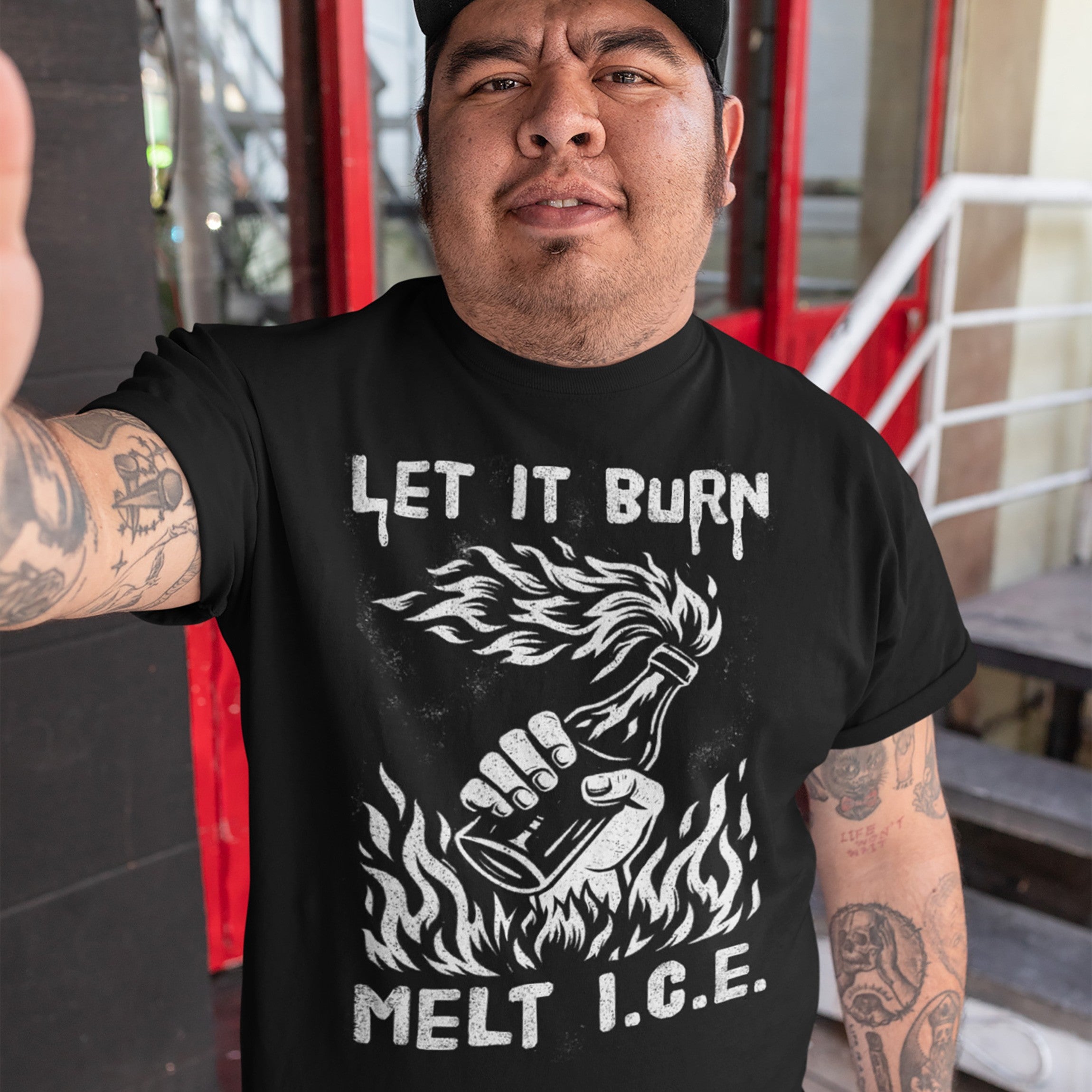 Person wearing the Let It Burn / Melt ICE T-shirt, featuring a Molotov cocktail graphic and bold text. Visible tattoos on forearms, set against an outdoor background with a red doorframe and metal stairs.