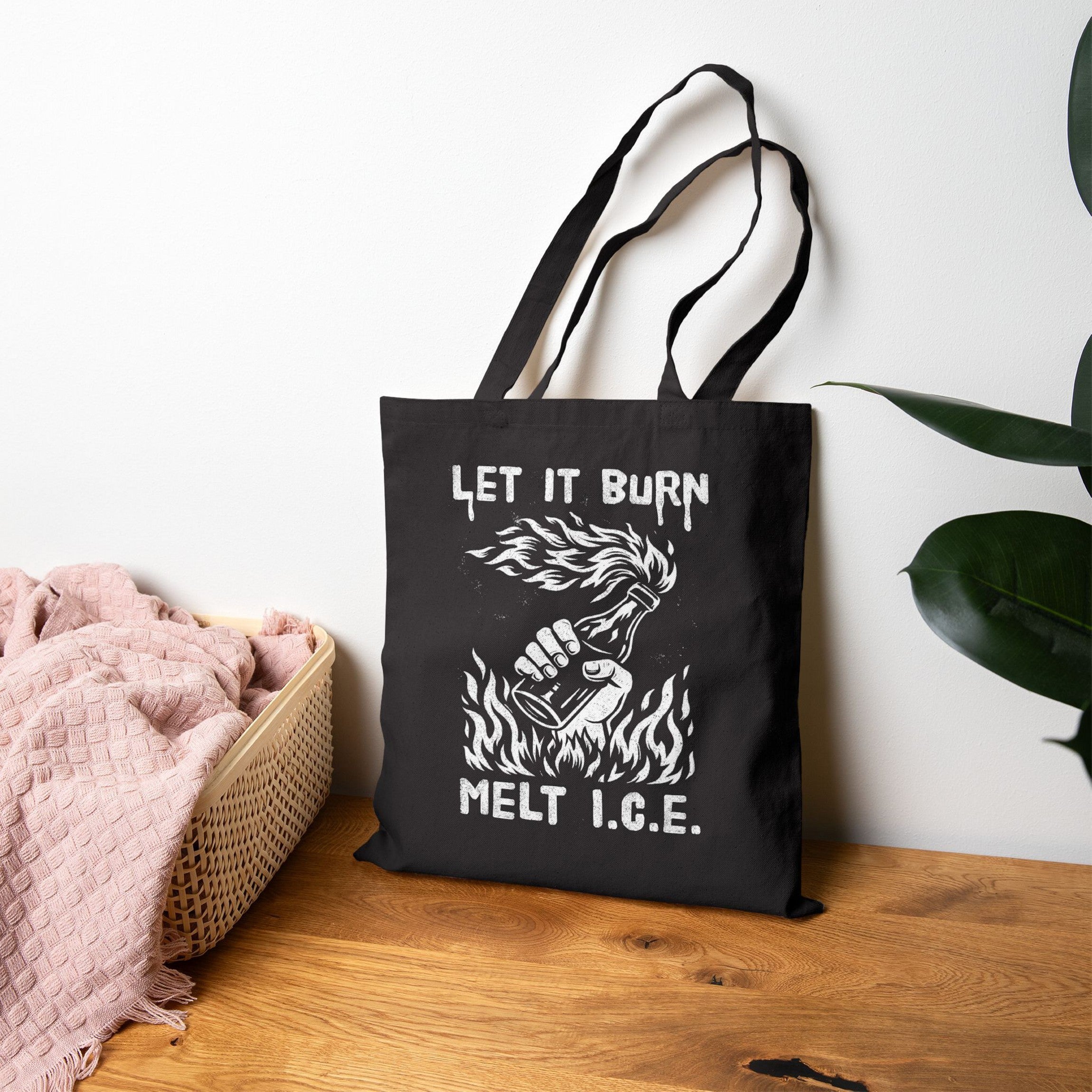 Let It Burn / Melt ICE Tote Bag with Molotov-cocktail graphic and slogan, on a wooden tabletop beside a basket and plant, symbolizes anti-fascist protest and immigrant rights advocacy.