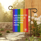 Custom Hate Has No Home Here Pride Garden Flag displayed on a black metal stand near a stone path, surrounded by white flowering shrubs, conveying a powerful message of inclusivity and equality.