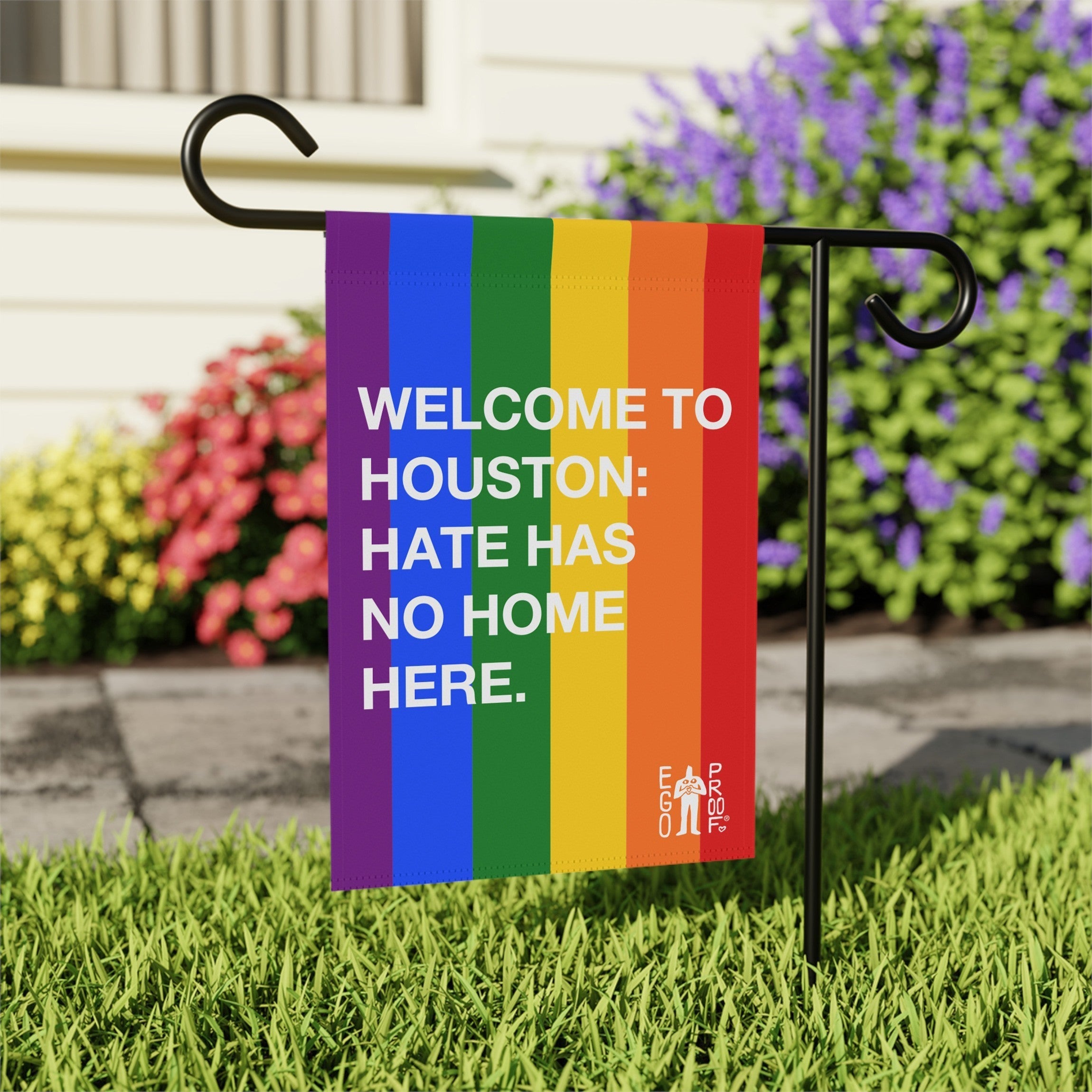 Custom Hate Has No Home Here Pride Garden Flag with rainbow stripes and bold white text, mounted on a black metal stand in a yard, promoting inclusivity and equality.