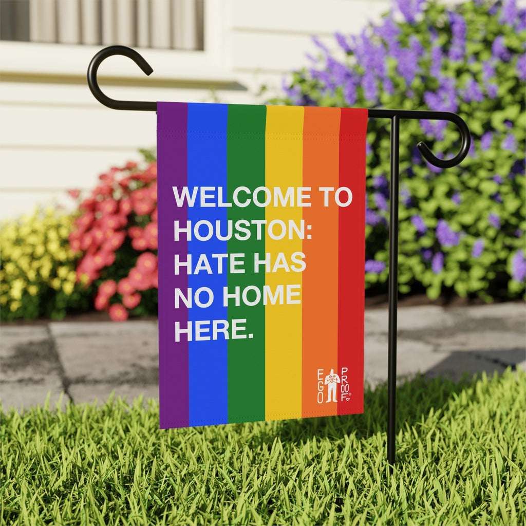 Custom Hate Has No Home Here Pride Garden Flag with rainbow stripes and bold white text, mounted on a black metal stand in a yard, promoting inclusivity and equality.