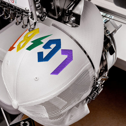 White LGBTQ Rocks Hat with rainbow embroidery being stitched on a mesh trucker hat showcasing pride and rock style.