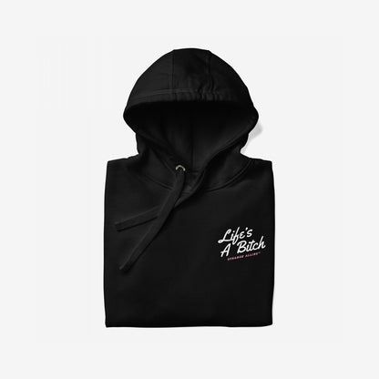 Black pullover hoodie titled Life's A Bitch Hoodie & Sweatshirt with a white slogan and pink text, embodying unapologetic realism. Hood and drawstrings visible, laid on a light background.