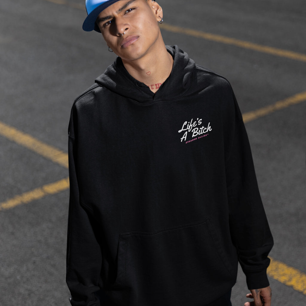 Person wearing a black Life's A Bitch hoodie with pink and white text, embodying a bold, unapologetic statement. The hoodie features a front pouch pocket and complements their blue cap and accessories.