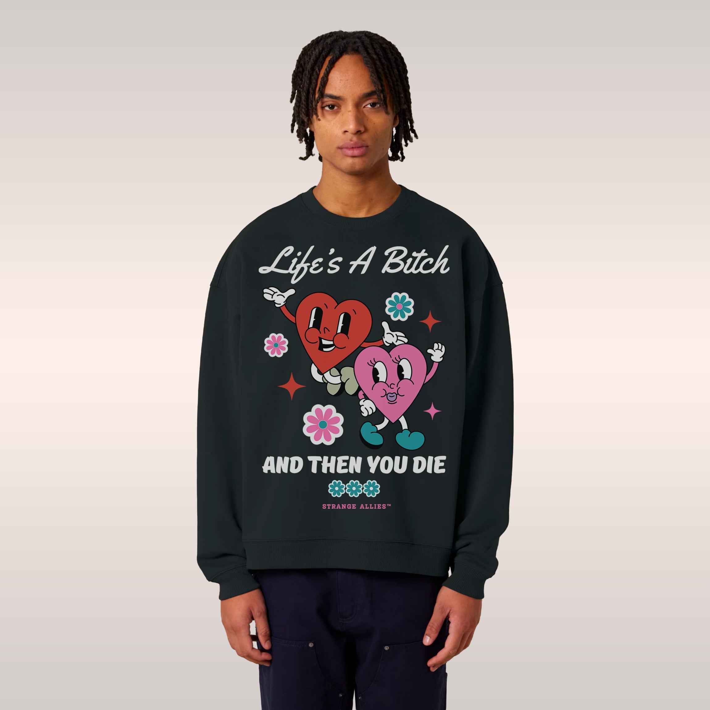 Young man on a ledge wearing a Life's A Bitch Shirt—black sweatshirt with cartoon hearts, flowers, stars, and bold text. A