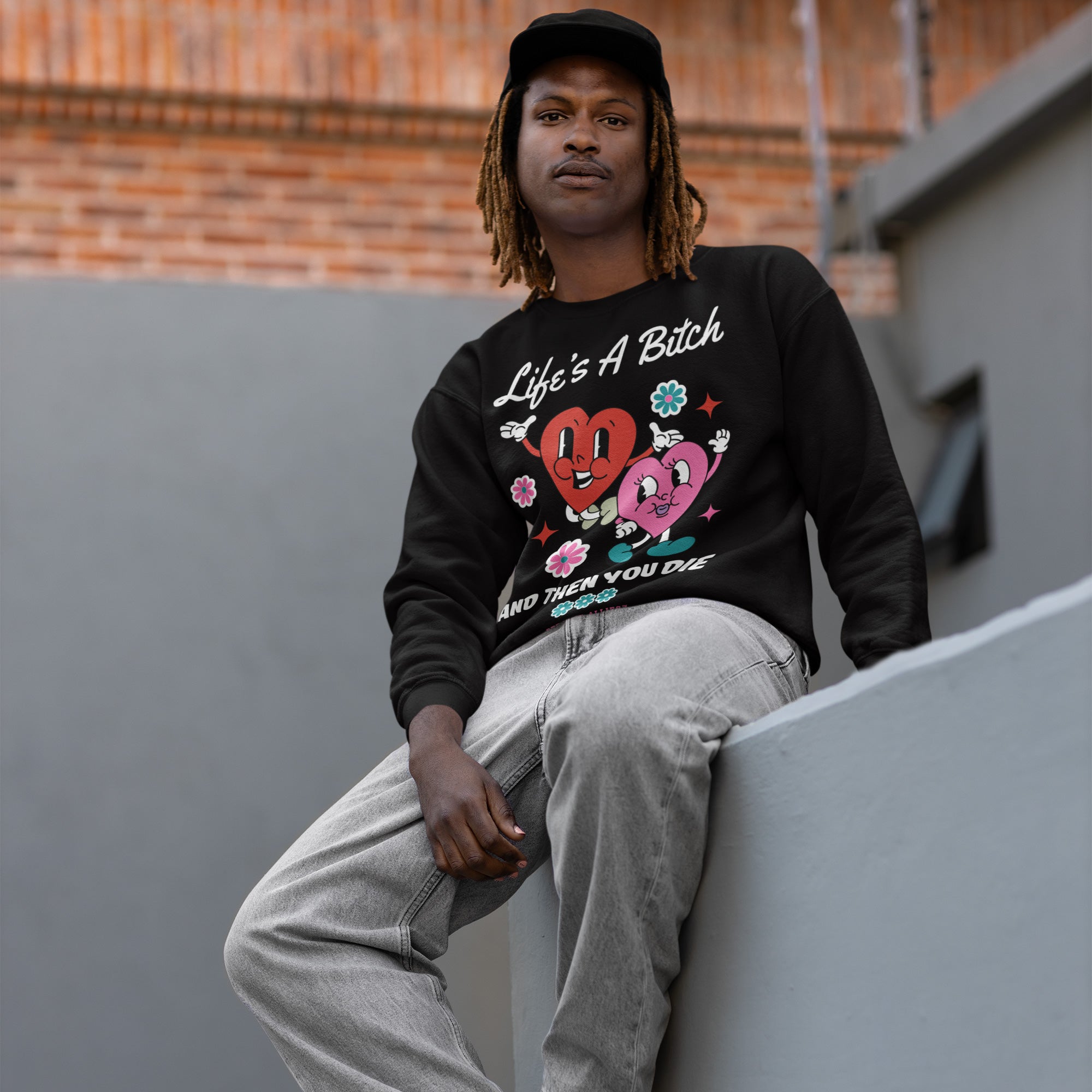 Young man on a ledge wearing a Life's A Bitch Shirt—black sweatshirt with cartoon hearts, flowers, stars, and bold text. A statement for those embracing life's truths.