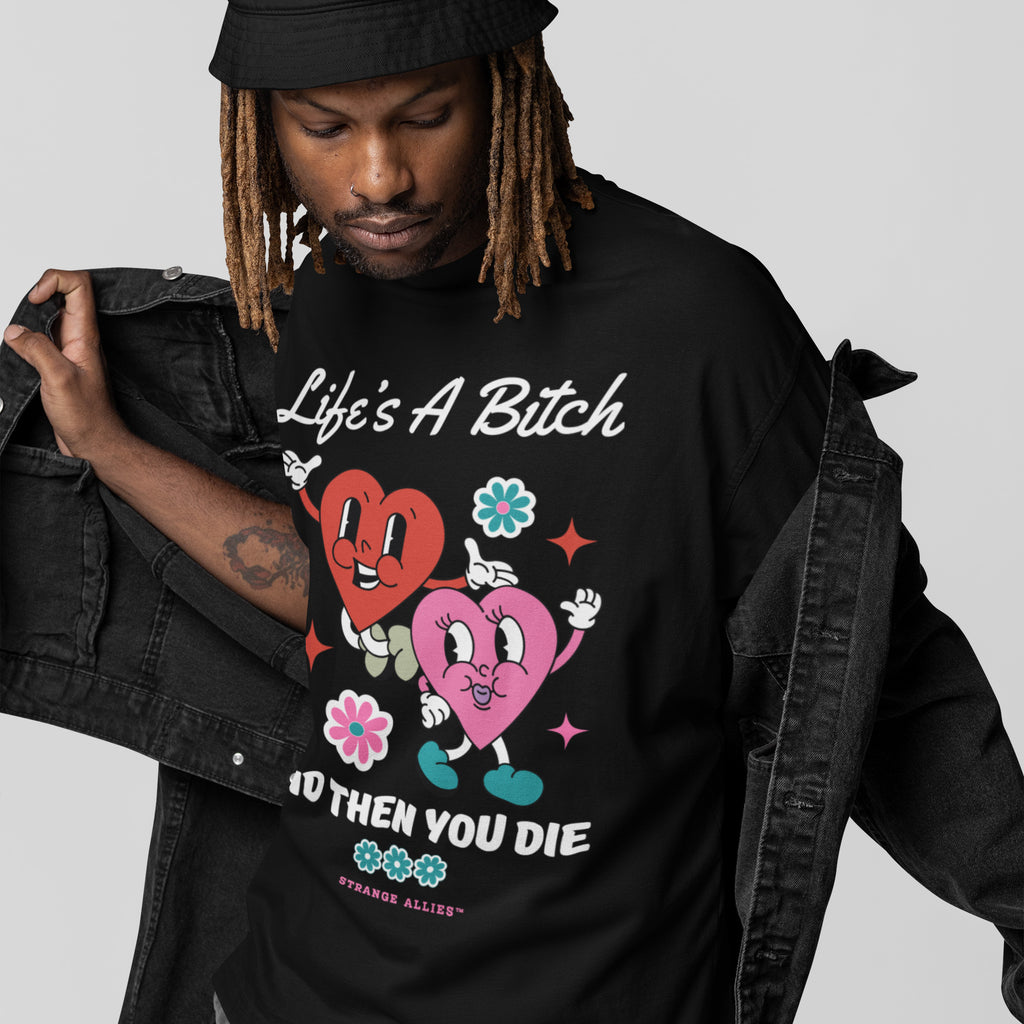 Person wearing a black bucket hat and denim jacket reveals the Life's A Bitch Shirt, featuring cartoon hearts, flowers, and bold text, embodying a candid, unapologetic message.