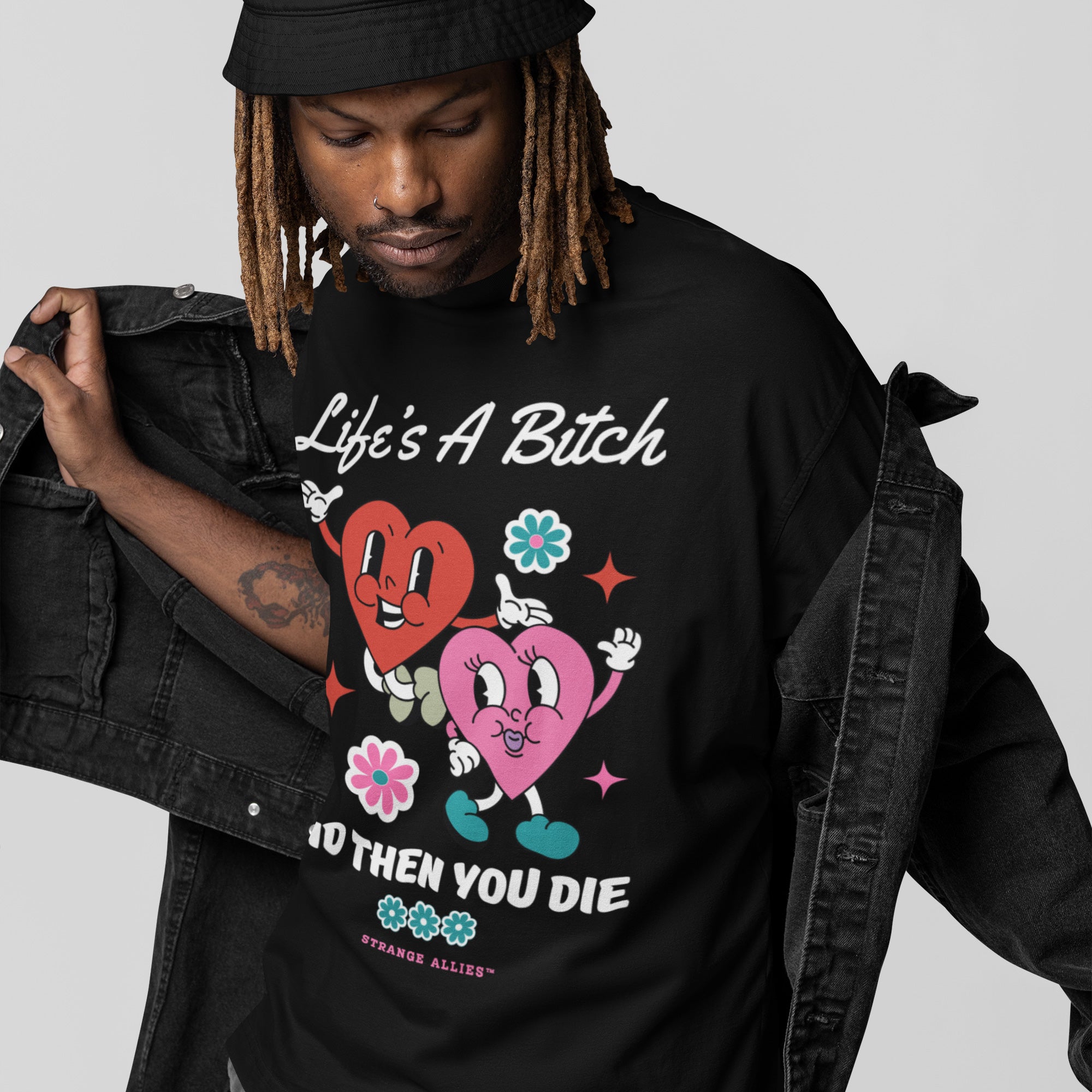 Person wearing a black bucket hat and denim jacket reveals the Life's A Bitch Shirt, featuring cartoon hearts, flowers, and bold text, embodying a candid, unapologetic message.