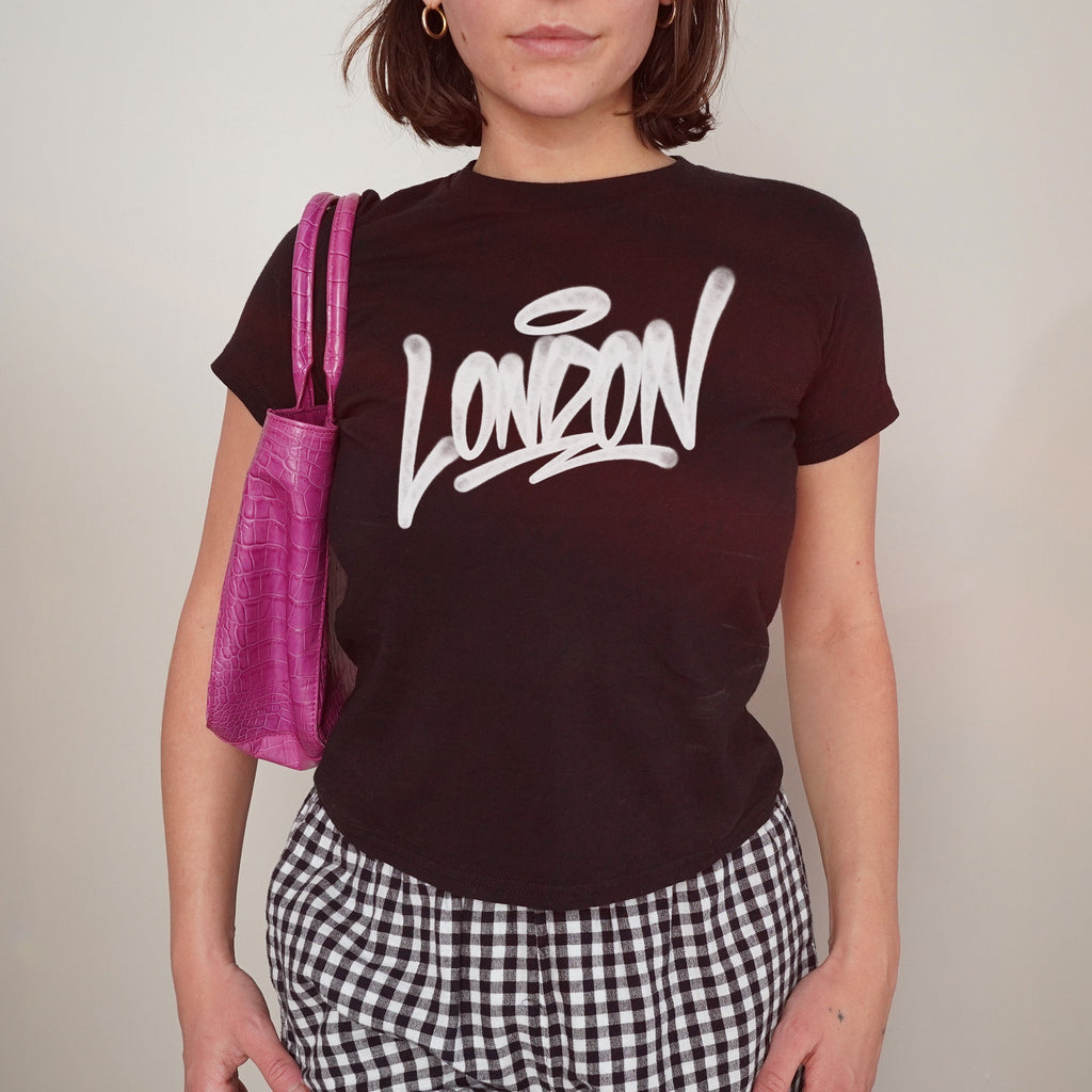 Person wearing the London Handstyle Women's Baby Tee, showcasing a graffiti-style London design in white. Paired with gingham pants and a fuchsia croc-embossed bag, embodying a bold, retro aesthetic.
