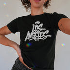 Person wearing the Los Angeles Handstyle Women's Baby Tee, featuring a white graffiti-style logo across the chest, with iridescent reflections highlighting the design. The tee embodies a bold, retro Y2K vibe.