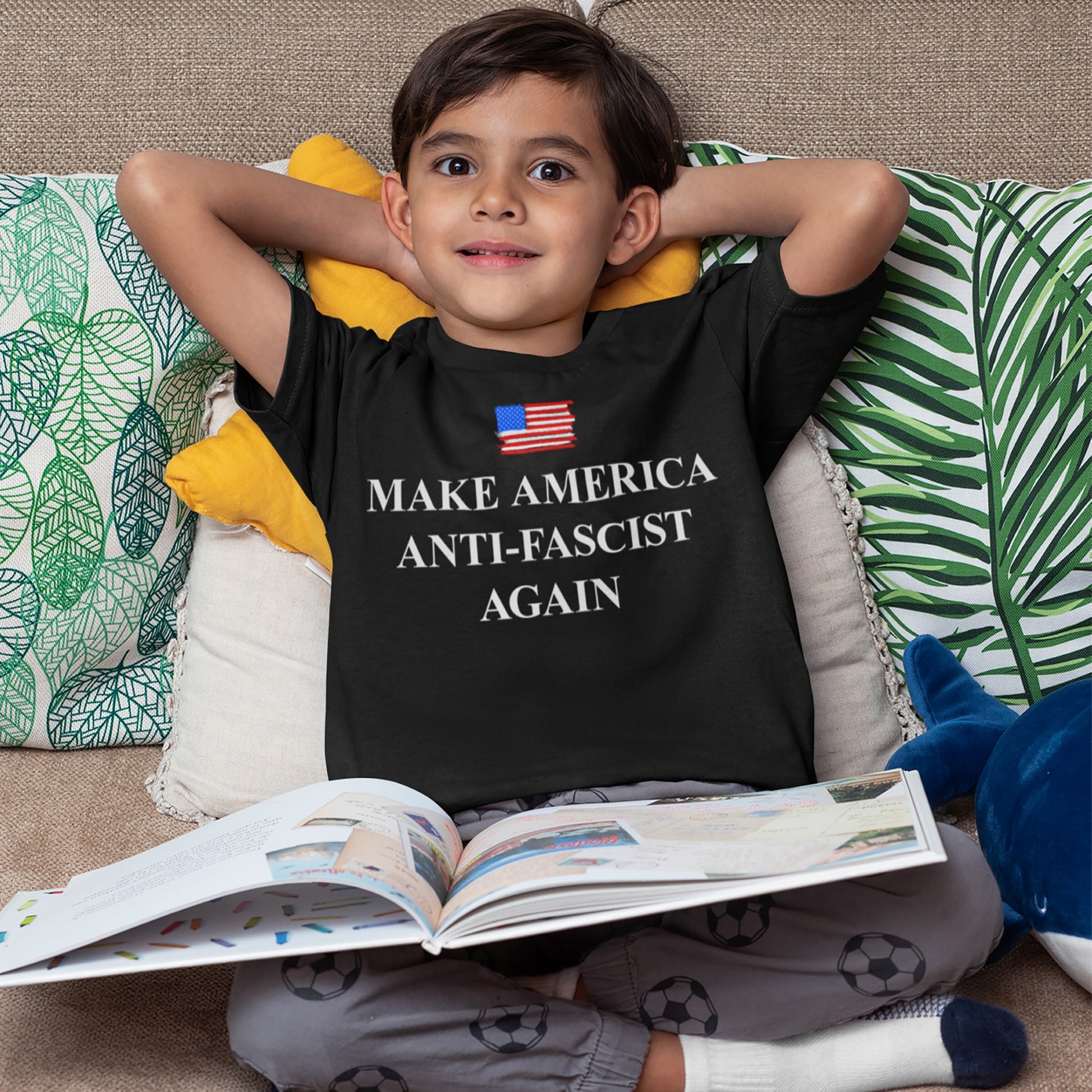 Young boy on a couch with a Make America Anti-Fascist Again T-shirt, featuring a small U.S. flag and bold serif slogan, holding an open picture book.