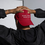 Person wears a Make America Rage Again Hat, bright red with white embroidery, under black jacket. Their hands rest on the cap, showcasing tattoos and a hoop earring, embodying a bold protest spirit.