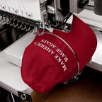 Red Make America Rage Again hat being embroidered on an industrial machine, showcasing anti-fascist slogan in bold white letters, reflecting a strong protest message against authoritarianism.