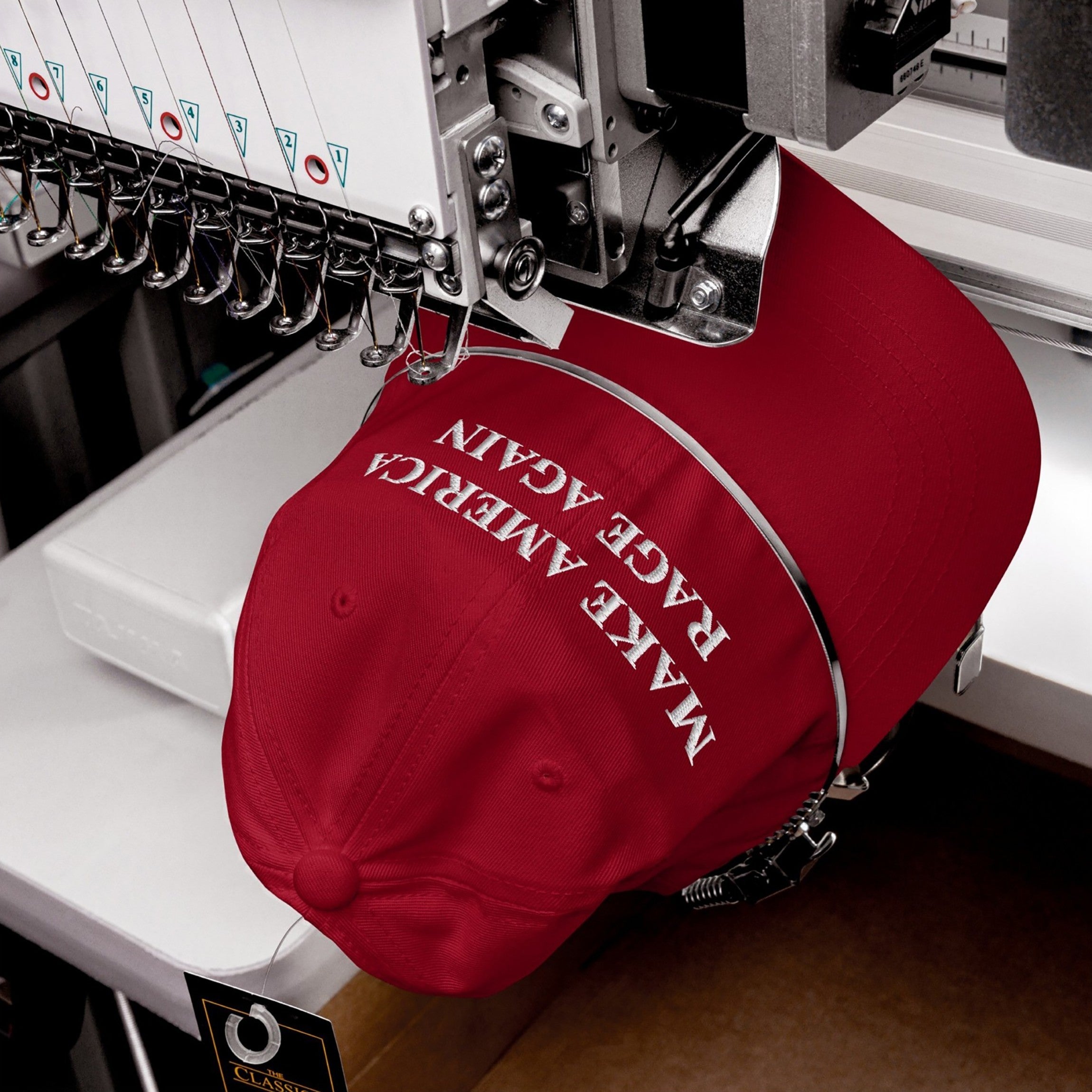 Red Make America Rage Again hat being embroidered on an industrial machine, showcasing anti-fascist slogan in bold white letters, reflecting a strong protest message against authoritarianism.