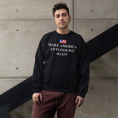 Young person wearing a black Make America Anti-Fascist Again sweatshirt with a small American flag, embodying anti-fascist values, standing confidently against a concrete wall with maroon pants and simple jewelry.