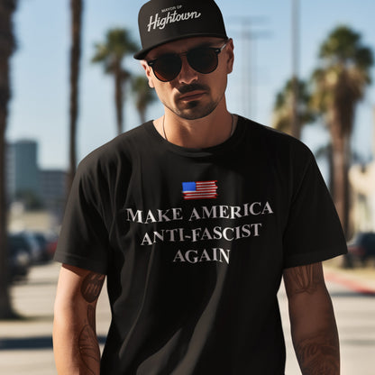 Man wearing a Make America Anti-Fascist Again T-shirt with a small American flag, black baseball cap, and sunglasses, standing outdoors. The shirt conveys a bold, progressive message against fascism.