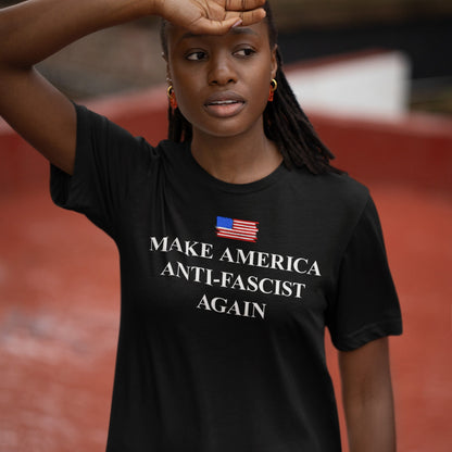 Young Black woman outdoors in a black Make America Anti-Fascist Again T-shirt with an American flag, embodying a strong anti-fascist message.
