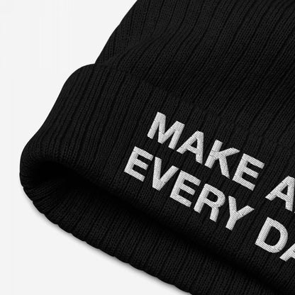 Close-up of the Make Art Every Day Hat, a black knit beanie with bold white embroidery reading “MAKE A” and “EVERY D” on the cuff, reflecting a creative mantra.