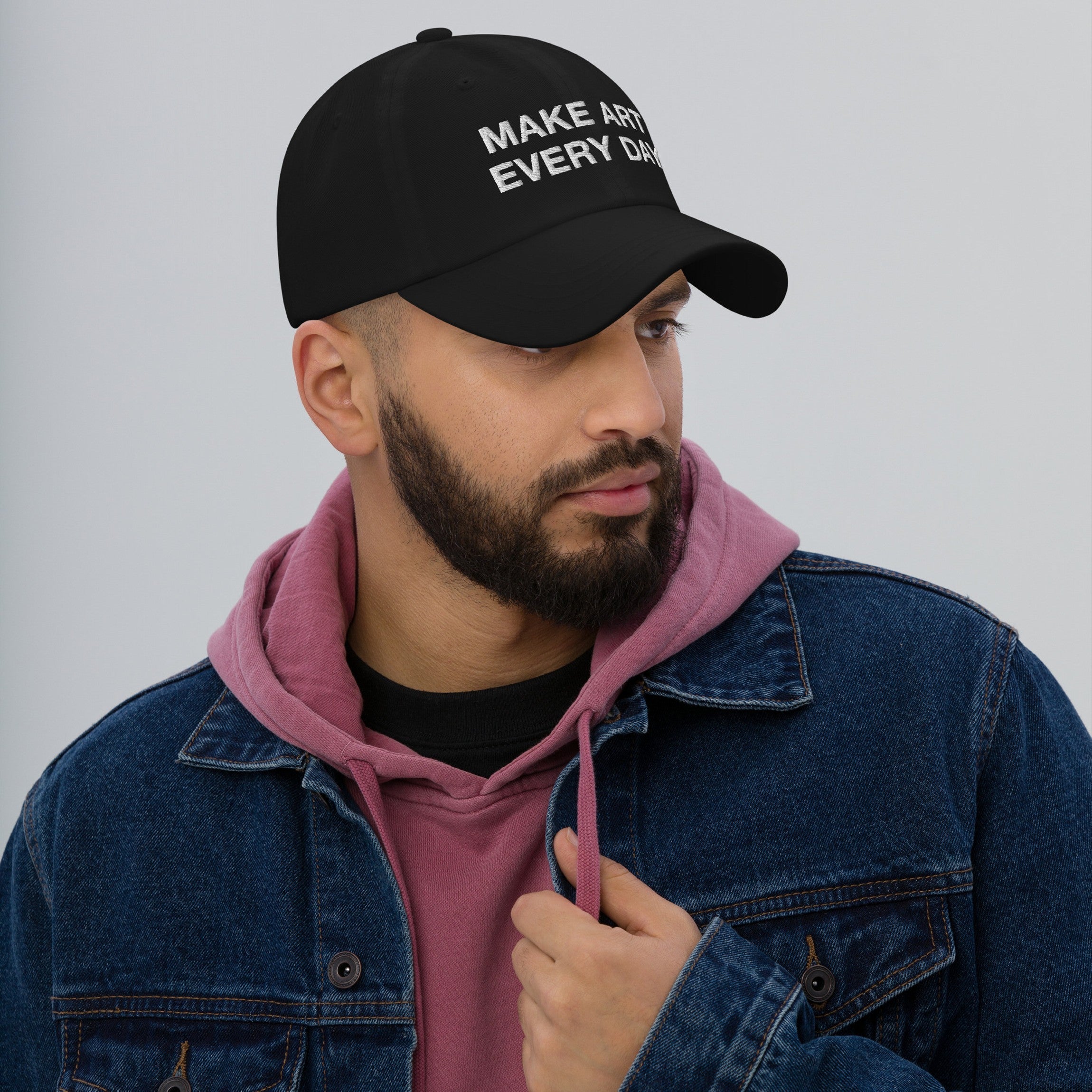 A young man wears the Make Art Every Day Hat, featuring bold embroidery. Styled with a denim jacket and hoodie, it embodies creativity and individuality, echoing a commitment to artistic expression.
