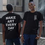 Two young adults showcase the Make Art Every Day T-shirt. One, facing backward, displays the bold back print, while the other, facing forward, highlights the understated front design, embodying creative street style.