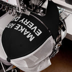 Close-up of Make Art Every Day Hat being embroidered on a commercial machine, showcasing bold white lettering on its black front panel, ideal for creative expression and comfortable wear during artistic endeavors.
