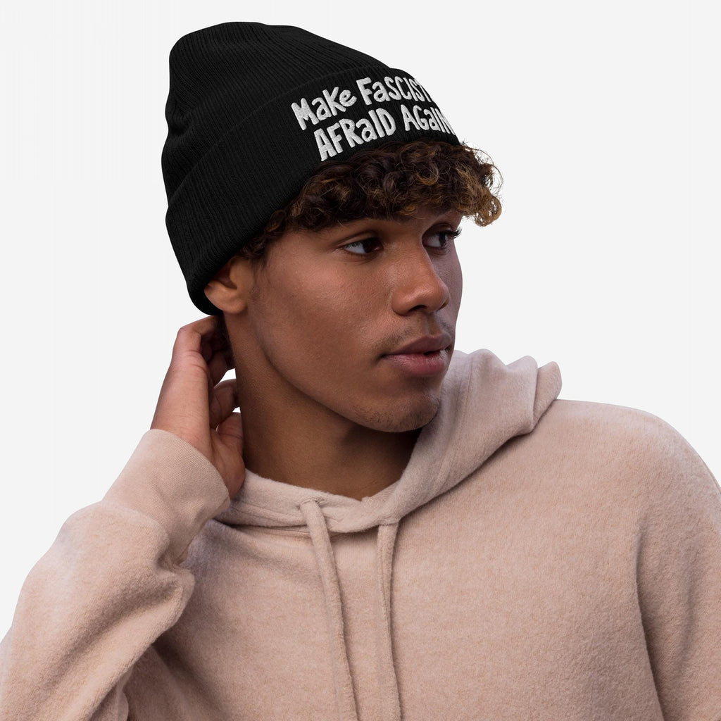 Young man in a light-tan hoodie wears the Make Fascists Afraid Again Hat, a black beanie with white embroidered slogan, embodying everyday resistance and defiance against fascism.