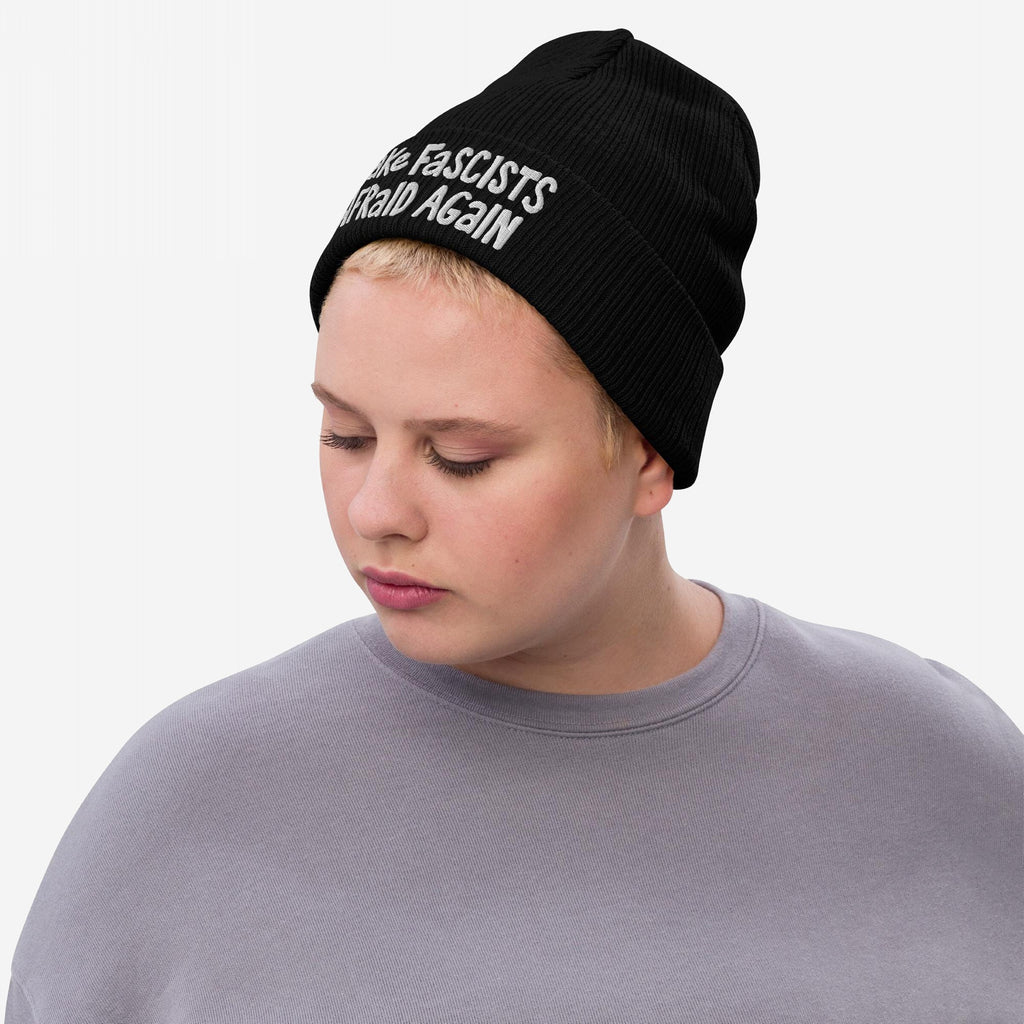 A young adult wears the Make Fascists Afraid Again hat, showcasing its bold slogan in raw, hand-drawn embroidery. The black knit beanie complements their understated gray top, embodying everyday resistance.