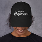 Person smiling broadly, wearing a black Mayor of Hightown baseball cap, signifying cannabis culture pride and humor, with dark curly hair peeking out, embodying a carefree, laid-back attitude.