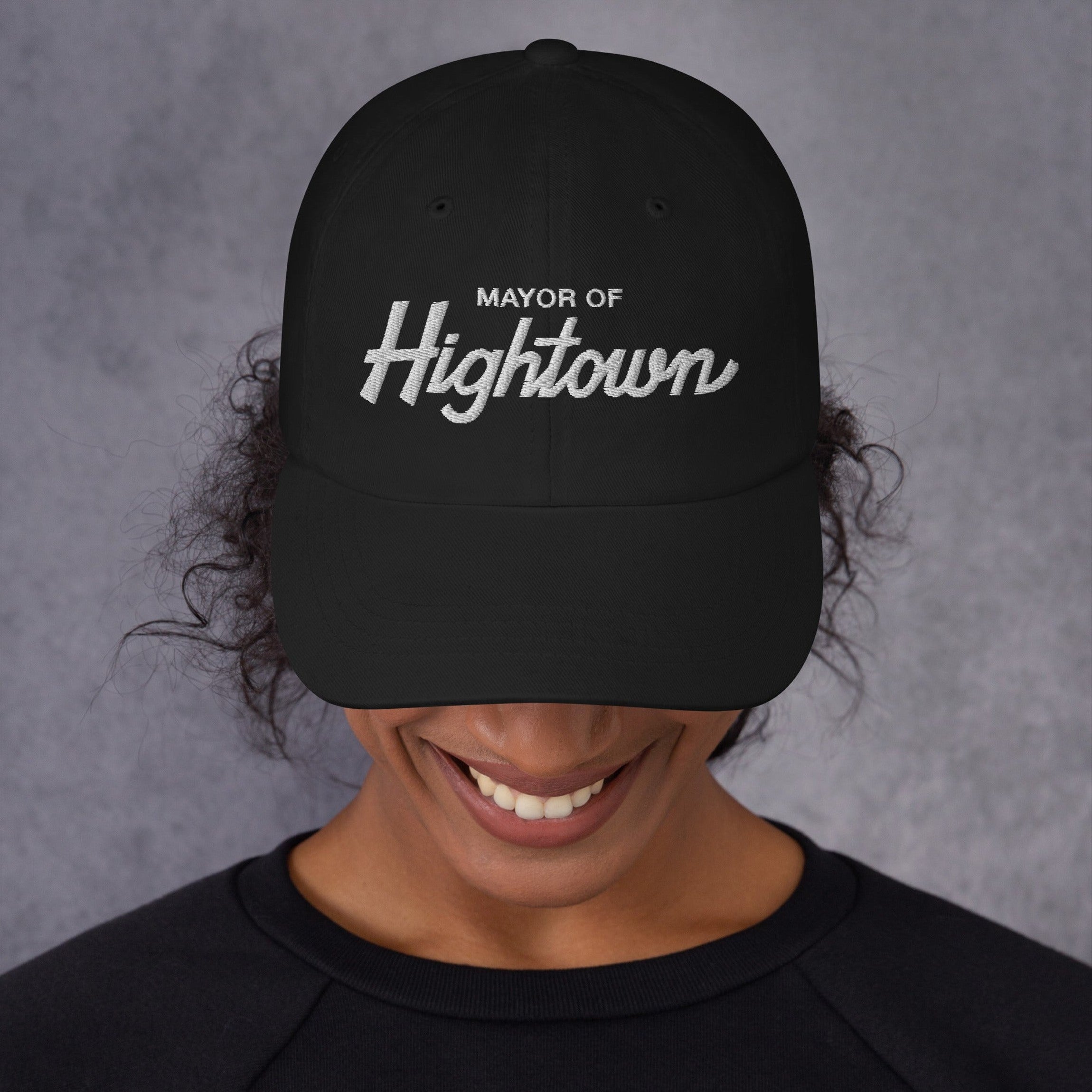 Person smiling broadly, wearing a black Mayor of Hightown baseball cap, signifying cannabis culture pride and humor, with dark curly hair peeking out, embodying a carefree, laid-back attitude.