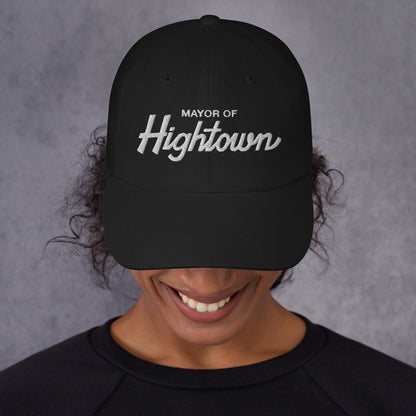 Person smiling broadly, wearing a black Mayor of Hightown baseball cap, signifying cannabis culture pride and humor, with dark curly hair peeking out, embodying a carefree, laid-back attitude.