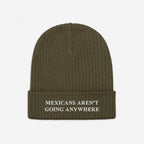 The Mexicans Aren’t Going Anywhere Hat features an olive-green rib-knit beanie with a folded cuff, embroidered with a bold, white message advocating pride and resilience against racism and anti-immigrant sentiments.