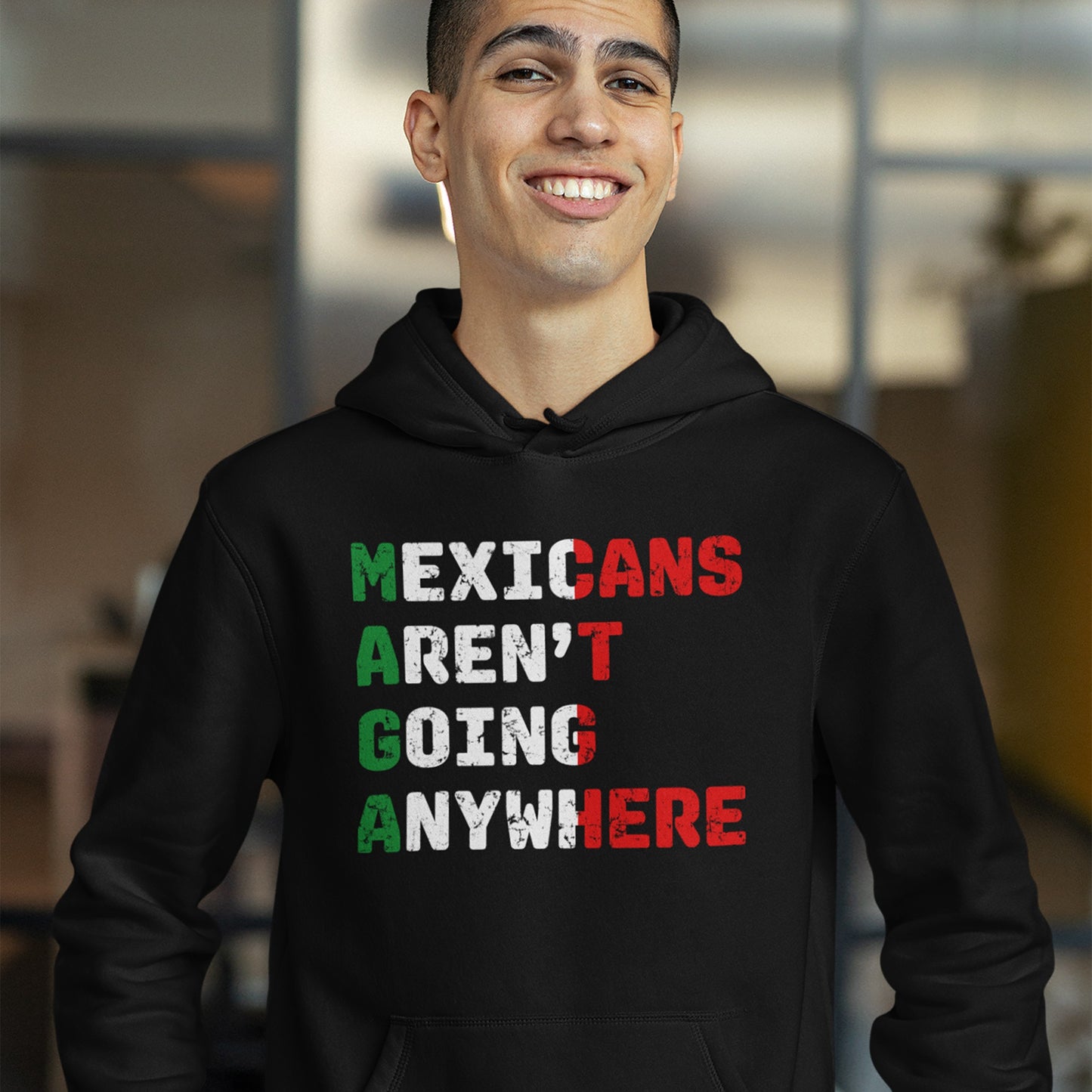 A young man wears a black hoodie featuring the message “Mexicans Aren’t Going Anywhere” vertically styled in Mexican flag colors, symbolizing pride and resistance.