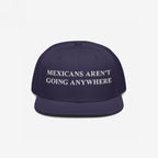 Navy snap-back cap with white embroidered Mexicans Aren’t Going Anywhere text, embodying pride and resistance against racism, available in multiple styles. Supports Immigrant Legal Resource Center.