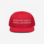 Red snap-back baseball cap with white embroidered text, “Mexicans Aren’t Going Anywhere,” symbolizing pride and resistance against racism and anti-immigrant sentiment. Supports Immigrant Legal Resource Center (ILRC).