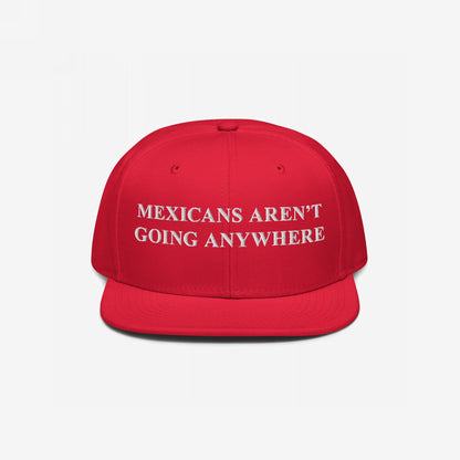 Red snap-back baseball cap with white embroidered text, “Mexicans Aren’t Going Anywhere,” symbolizing pride and resistance against racism and anti-immigrant sentiment. Supports Immigrant Legal Resource Center (ILRC).