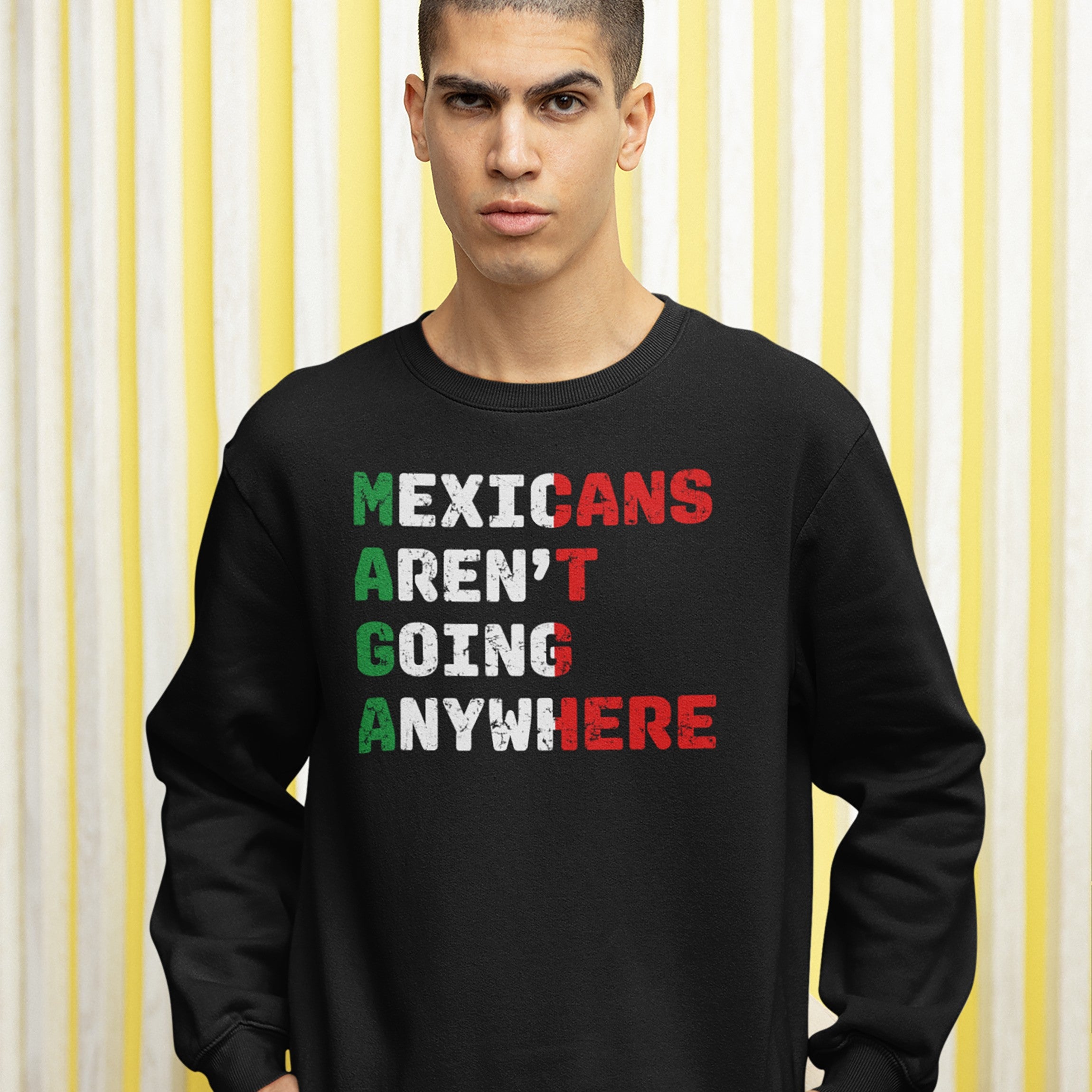 Young man wearing the Mexicans Aren’t Going Anywhere sweatshirt with bold block letters. The design creatively highlights MAGA, symbolizing defiance and pride, amidst a simple striped backdrop.