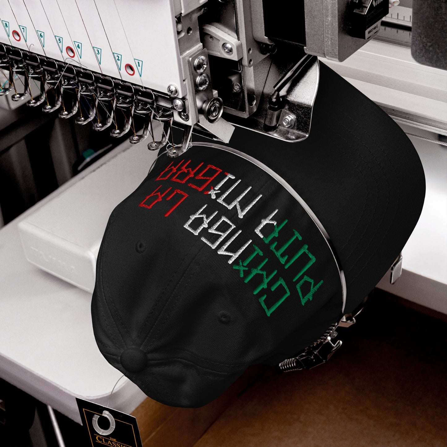 The Chinga La Migra Mexico Hat is being embroidered with bold lettering on a computerized machine, featuring multi-colored threads representing the Mexican flag, and a “Classic” tag on the back strap.