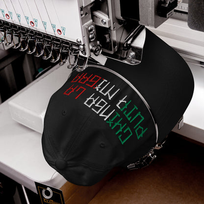 The Chinga La Migra Mexico Hat is being embroidered with bold lettering on a computerized machine, featuring multi-colored threads representing the Mexican flag, and a “Classic” tag on the back strap.