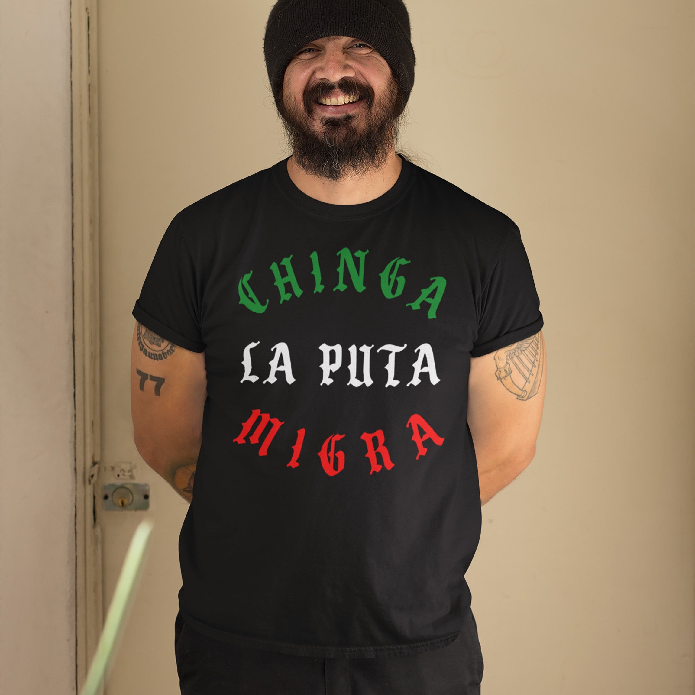 A bearded man wears the Chinga La Migra Mexico T-shirt, featuring bold Mexican flag-colored text, supporting immigrant rights, with tattoos visible on his arms. He's smiling, standing against a light background.