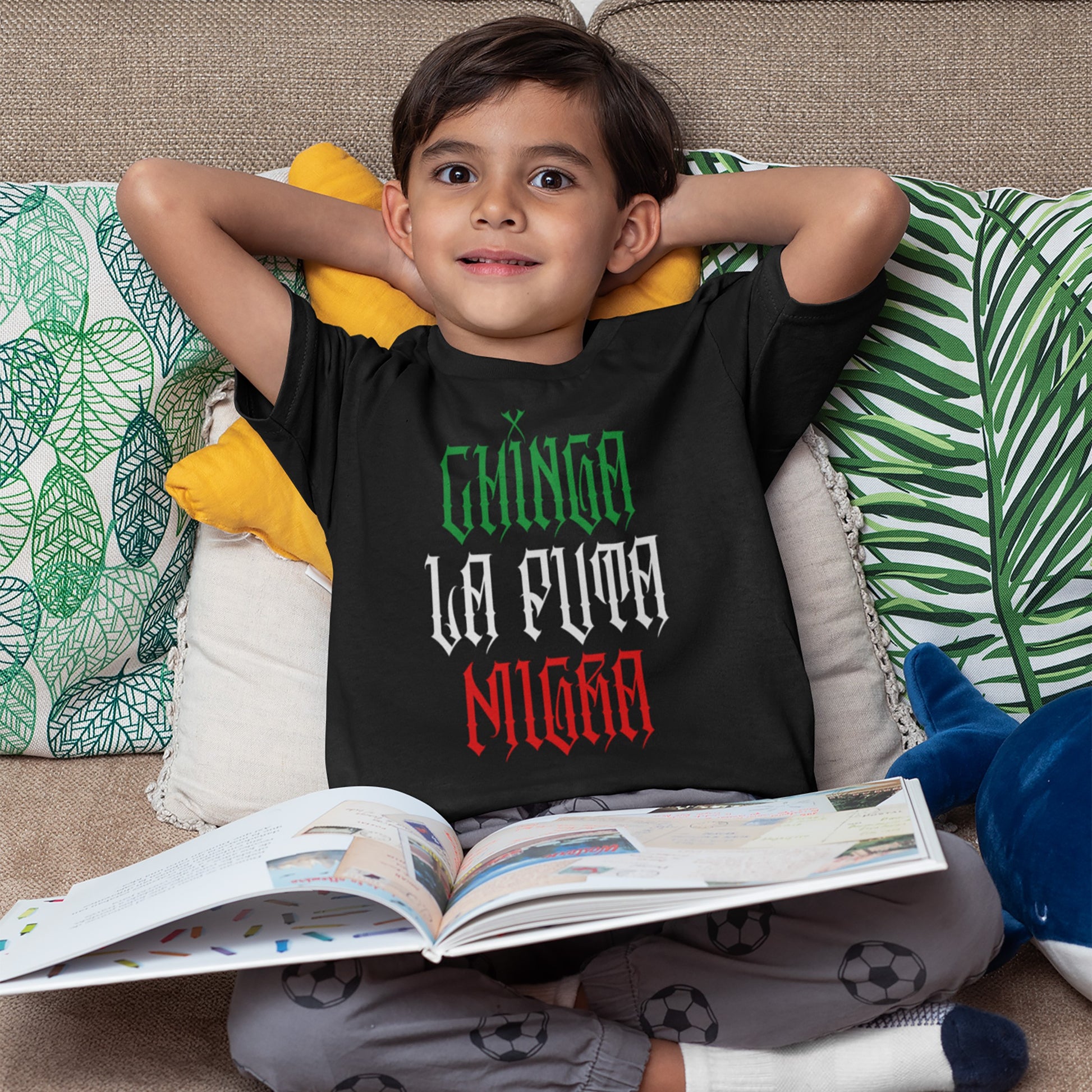 Young boy in gray pajama pants on couch, wearing a Chinga La Migra Mexico T-shirt with Mexican flag colors, symbolizing cultural pride and resistance against anti-immigrant systems. Blue shark plush beside him.