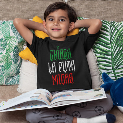 Young boy in gray pajama pants on couch, wearing a Chinga La Migra Mexico T-shirt with Mexican flag colors, symbolizing cultural pride and resistance against anti-immigrant systems. Blue shark plush beside him.