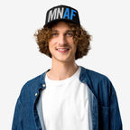 Young man wearing a Minnesota As Fuck Anti-Fascist Hat, featuring bold block letters, embodies pro-equality spirit with a black cap, open denim shirt, and a confident smile.