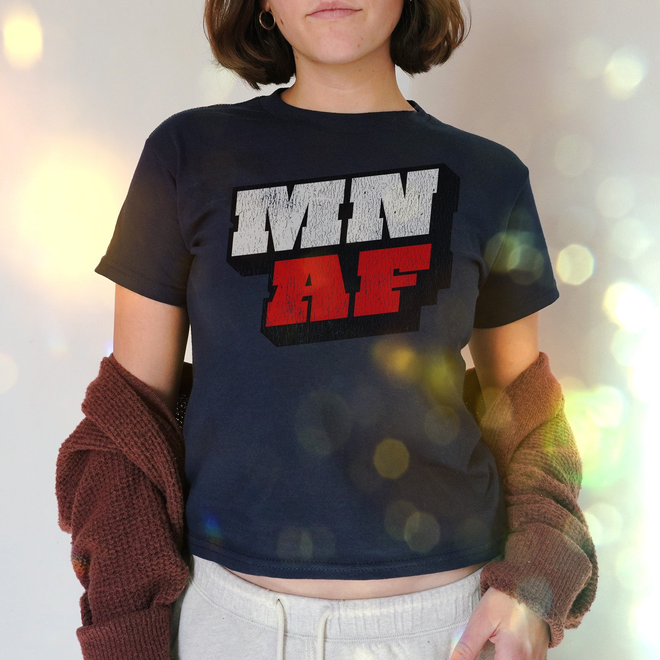 Minnesota As Fuck Anti-Fascist Women's Baby Tee in navy, featuring bold MN AF lettering. Styled with a rust cardigan and gray lounge pants, embodying defiant, retro Y2K vibes for a socially conscious statement.