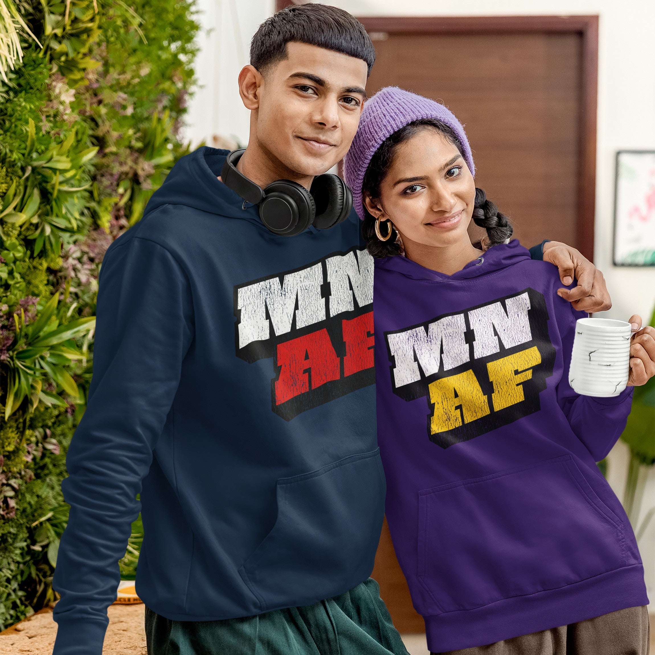 Two young adults indoors wearing Minnesota As Fuck Anti-Fascist Hoodies; left in navy blue, right in purple with matching beanie, symbolizing equality and anti-fascism. Both smile softly, one holding a mug.