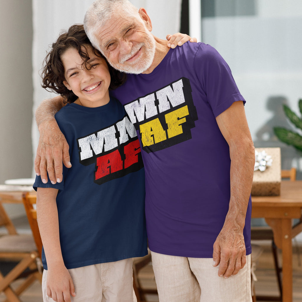 Older man and young boy, smiling and hugging, each wearing a Minnesota As Fuck Anti-Fascist T-shirt, showcasing bold block lettering. A wooden table with a wrapped gift is visible behind them.