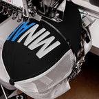 Industrial embroidery machine stitching MN AF on a black trucker cap, embodying the Minnesota As Fuck Anti-Fascist Hat's pro-equality and anti-fascism message with white and blue threads.