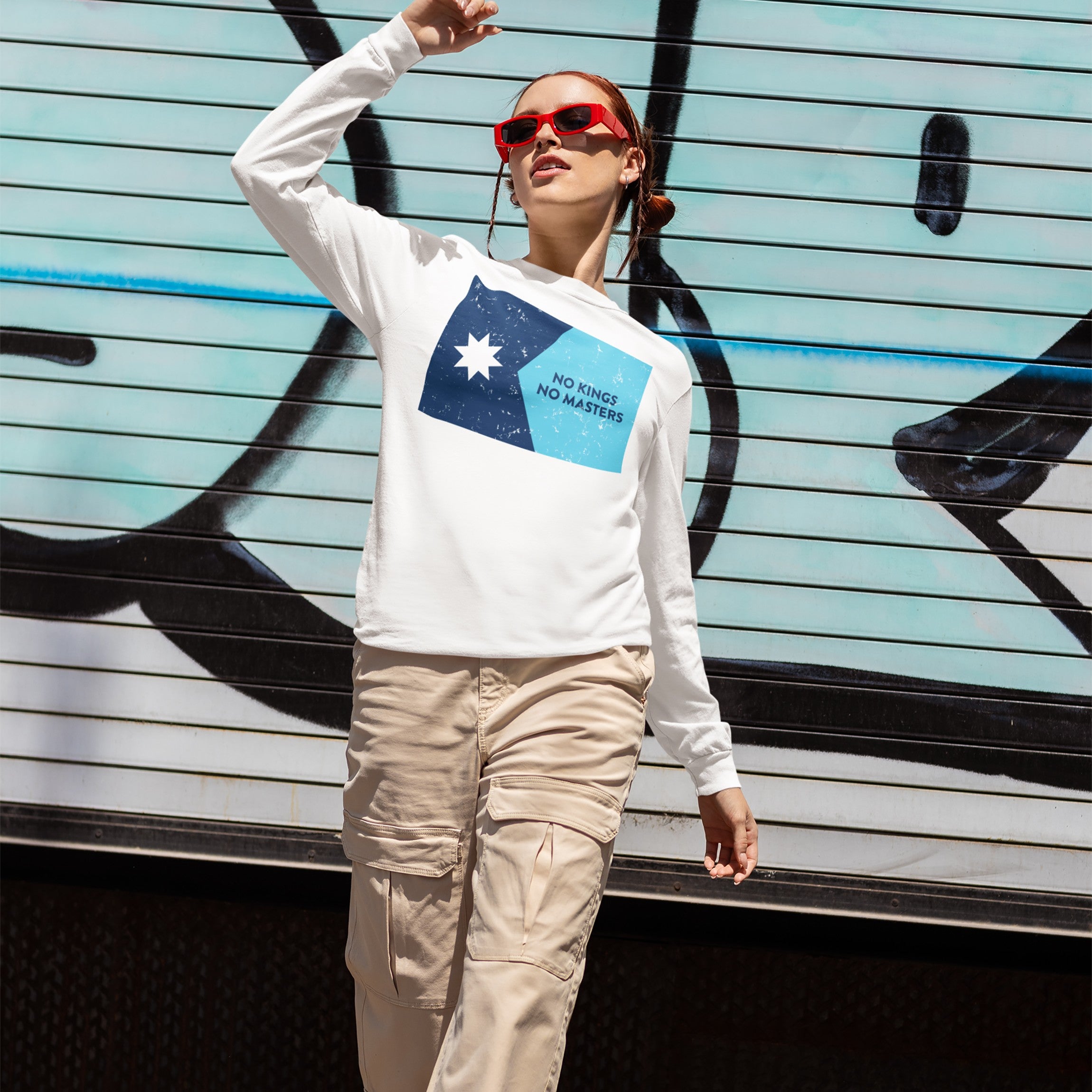 Person wearing the Minnesota No Kings No Masters Flag Shirt, featuring a two-tone blue graphic and slogan, stands before a graffiti backdrop, embodying a rebellious streetwear style against authoritarianism.