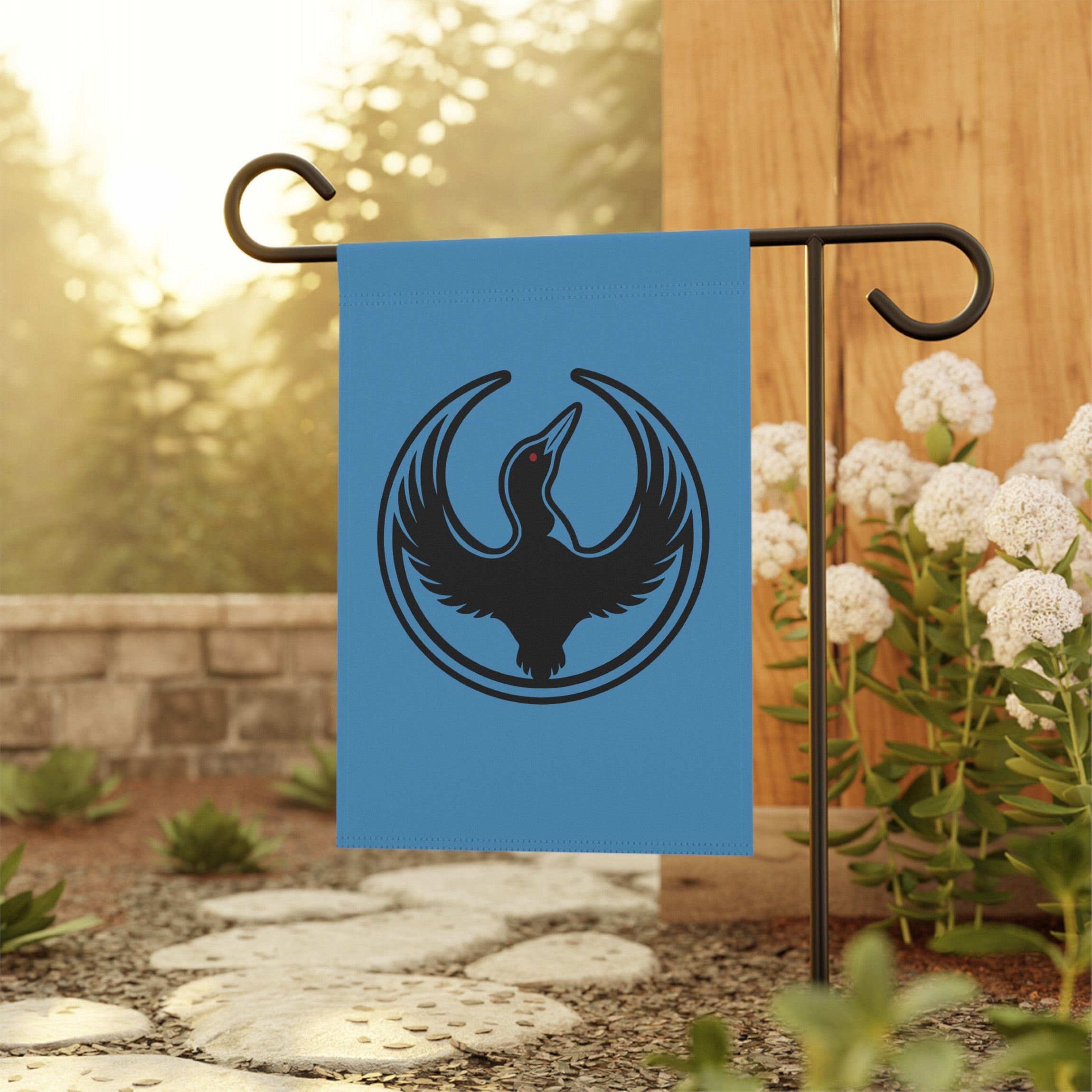 Minnesota Rebel Alliance Garden Flag features a stylized black loon on bright blue, displayed on a metal stand beside a flowerbed, embodying a stance for immigrant rights and equality.